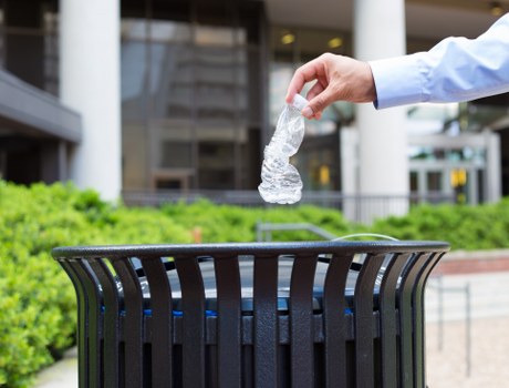 A person wearing a light blue long-sleeved shirt is seen discarding a crumpled clear plastic water bottle into a black outdoor rubbish bin with vertical slats, positioned on a paved surface in an urban environment. Behind the bin, there are low green shrubbery and blurred modern office buildings, indicating a contemporary commercial or residential area. The scene suggests waste disposal as part of routine rubbish removal services in Hampstead, with the focus on proper waste segregation of plastics. The image captures a moment of responsible littering, emphasizing the importance of organised rubbish collection in the local community, in line with services offered by Commercial Waste Removal Hampstead.