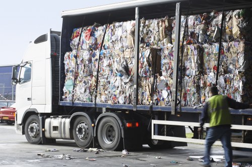 A large commercial rubbish collection truck parked on a paved surface, filled with tightly packed compressed waste materials consisting of mixed paper, cardboard, and other household waste, visible through the open side panels. The truck has a white cab with a modern design, and its black container is filled with a variety of crumpled and stacked rubbish that appears to include printed papers, cardboard boxes, and plastic packaging, with some loose waste scattered on the ground nearby. A person wearing a high-visibility vest and dark clothing is walking beside the truck, possibly a waste management worker, while a red vehicle is partially visible in the background. The scene is set outdoors, likely in an urban or commercial area, with bright natural lighting illuminating the vehicle and waste contents. The image exemplifies the type of heavy-duty rubbish removal services offered by Commercial Waste Removal Hampstead, serving properties in the Hampstead area or nearby postal codes.