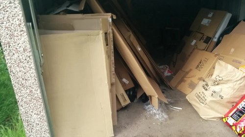 Inside a small, cluttered storage area or garage, there is a collection of various discarded items including flattened cardboard boxes, some partially torn or crumpled, and a large cream-colored wooden or MDF panel leaning against the left side. To the right, there are more cardboard boxes of different sizes, some with visible labels, and a crumpled plastic wrapping or packaging material lying among the debris. The background appears dark, with limited visibility of other objects, and the ground is concrete. The scene suggests a typical rubbish collection setup for waste removal services in Hampstead, suitable for a commercial or residential property, with the clutter indicating scheduled clearance or disposal of household or business waste, serviced by a professional waste removal company such as Commercial Waste Removal Hampstead.