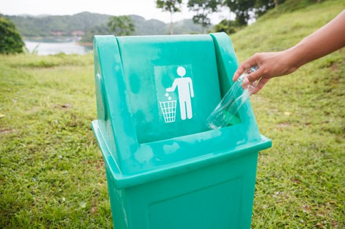 A person's hand is seen placing a clear plastic bottle into a bright turquoise recycling bin with a white icon of a person disposing of waste, situated outdoors on a grassy area. In the background, there are trees, a body of water, and a gently sloping green hillside, suggesting a park or natural environment near Hampstead. The bin is positioned on a paved or compacted surface, surrounded by well-maintained grass, with the scene illuminated by natural daylight. The image visually emphasizes waste disposal and recycling practices in a scenic outdoor setting, supporting environmentally conscious rubbish removal services offered by companies such as Commercial Waste Removal Hampstead in the local area.