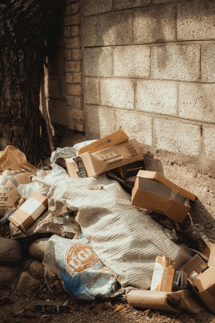 A collection of overflowing rubbish bins and scattered waste bags piled on a paved area in front of a commercial building, with various materials such as cardboard boxes, plastic bags, paper, and pack