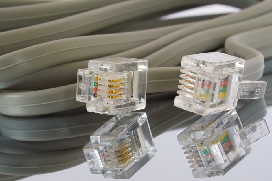 A close-up view of a coiled, light grey Ethernet cable with four transparent RJ45 connectors placed on a reflective surface. The connectors reveal gold-plated contact pins and small, colored wiring in